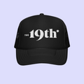 A black trucker-style cap with white text reading 'The 19th*' on the front, shown against a light lavender background.
