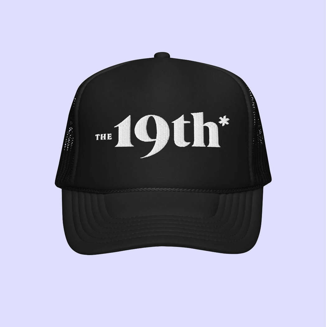 A black trucker-style cap with white text reading 'The 19th*' on the front, shown against a light lavender background.