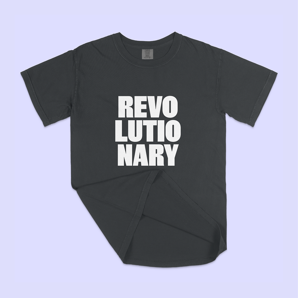A dark charcoal gray t-shirt with the word 'REVOLUTIONARY' printed in bold white capital letters across the chest, laid flat on a light lavender background.