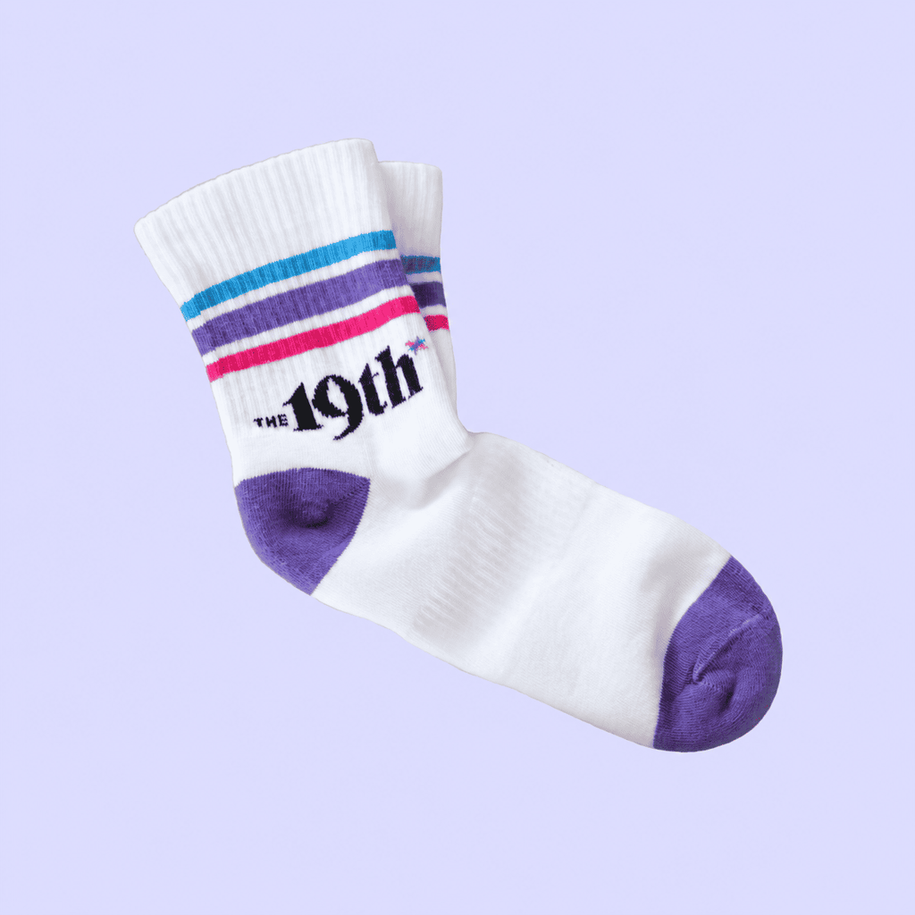 A pair of white crew socks with purple, blue, and pink stripes near the cuff, featuring the text 'THE 19th' in black. The toes and heels are solid purple.