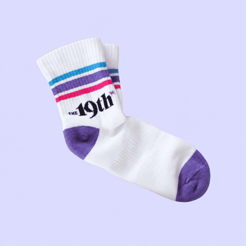 A pair of white crew socks with purple, blue, and pink stripes near the cuff, featuring the text 'THE 19th' in black. The toes and heels are solid purple.