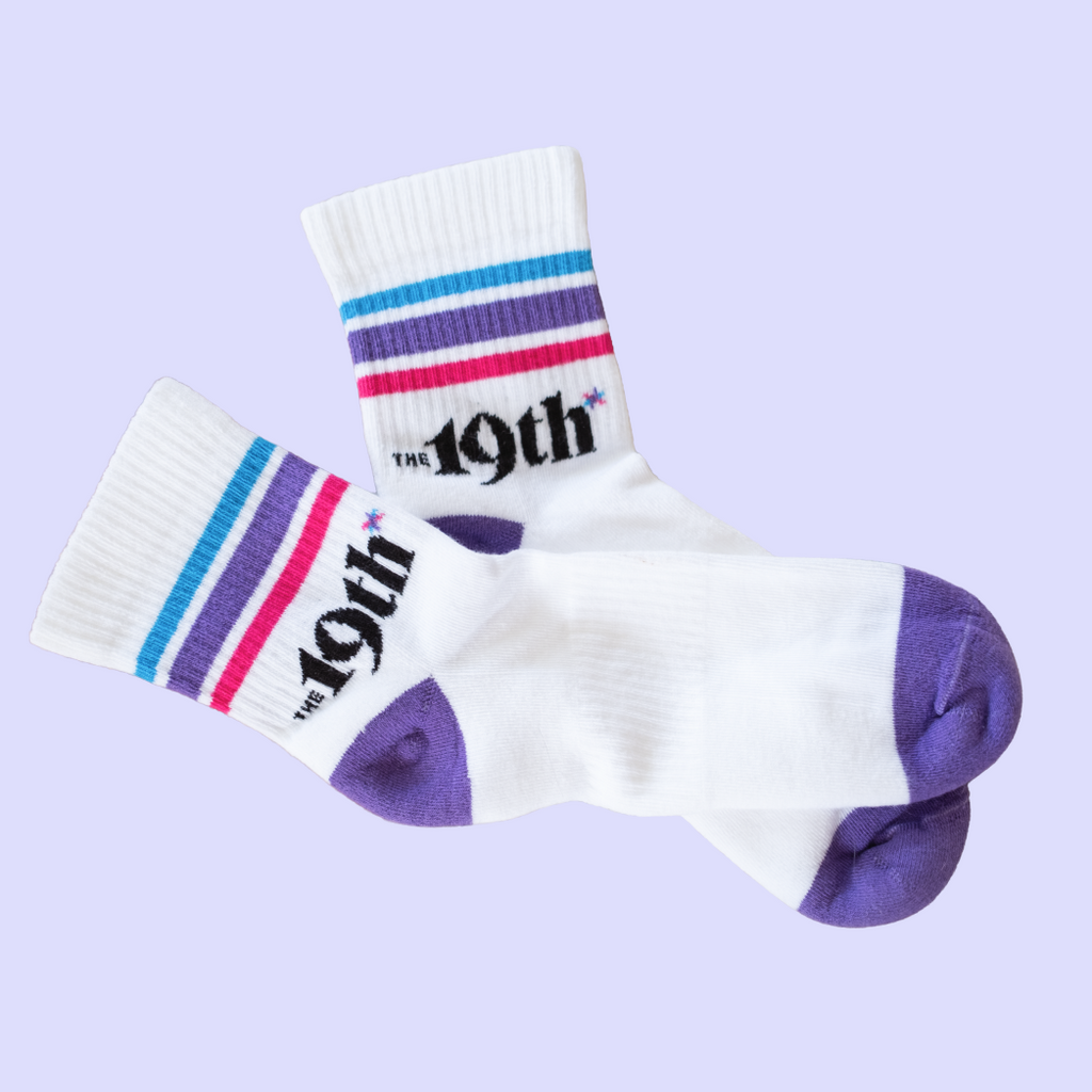 A pair of white crew socks with purple and blue stripes on the cuffs, pink stripes on one cuff, and purple toes and heels. The socks feature the text 'THE 19th' in a black font with a small asterisk element.